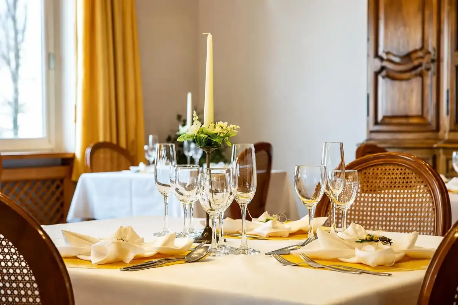 Elegante tavolo da pranzo apparecchiato con tovaglia bianca, bicchieri da vino, candele e fiori in una stanza luminosa.