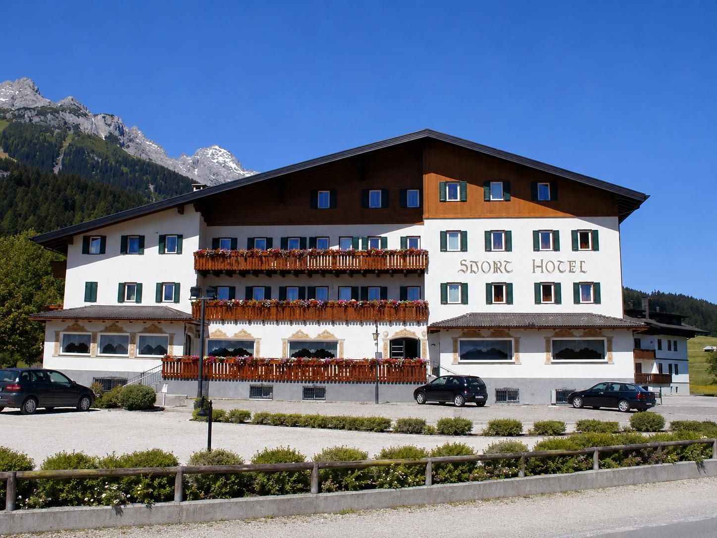 Hotel alpino con balconi in legno, auto parcheggiate e montagne sullo sfondo, sotto un cielo azzurro limpido.