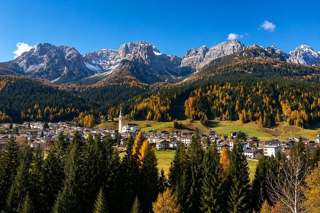 Villaggio di montagna con alberi autunnali sotto cime rocciose innevate, sotto un cielo azzurro e limpido.