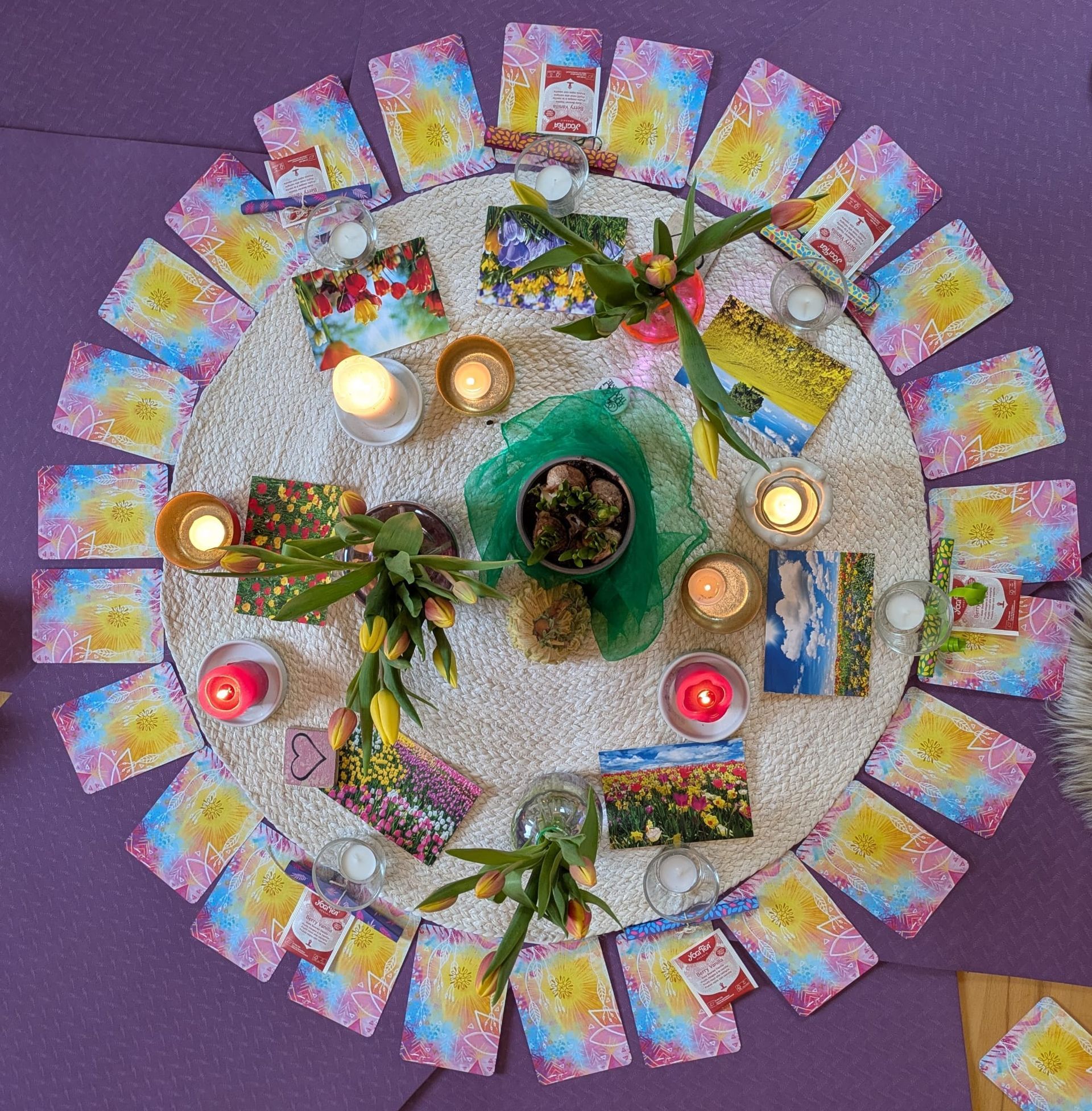 Eine kreisförmige Anordnung von Tarotkarten, Kerzen und gelben Tulpen auf einer weißen, strukturierten Matte vor einem violetten Hintergrund.