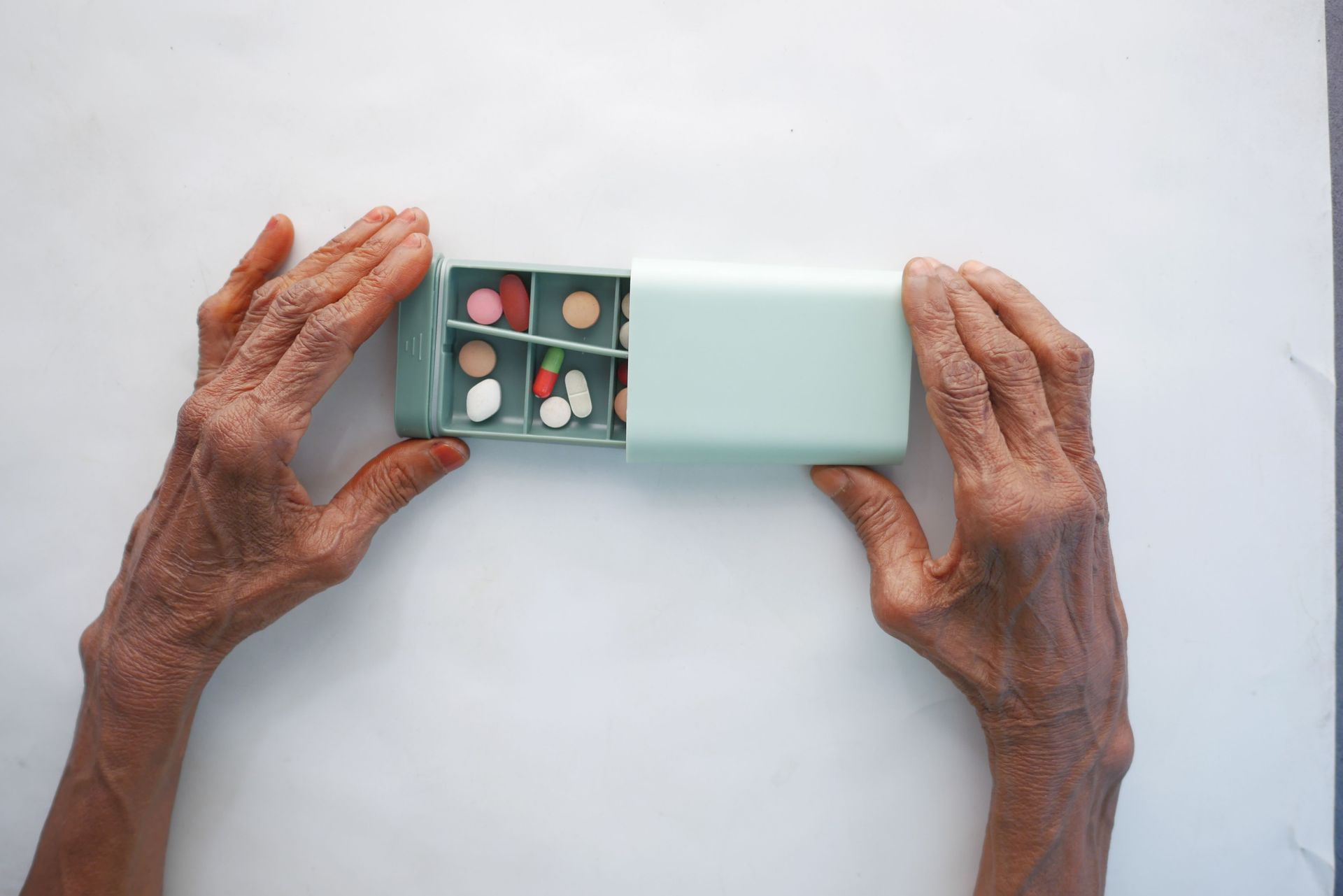 Hands opening a mint-green pill case with assorted tablets on a white surface