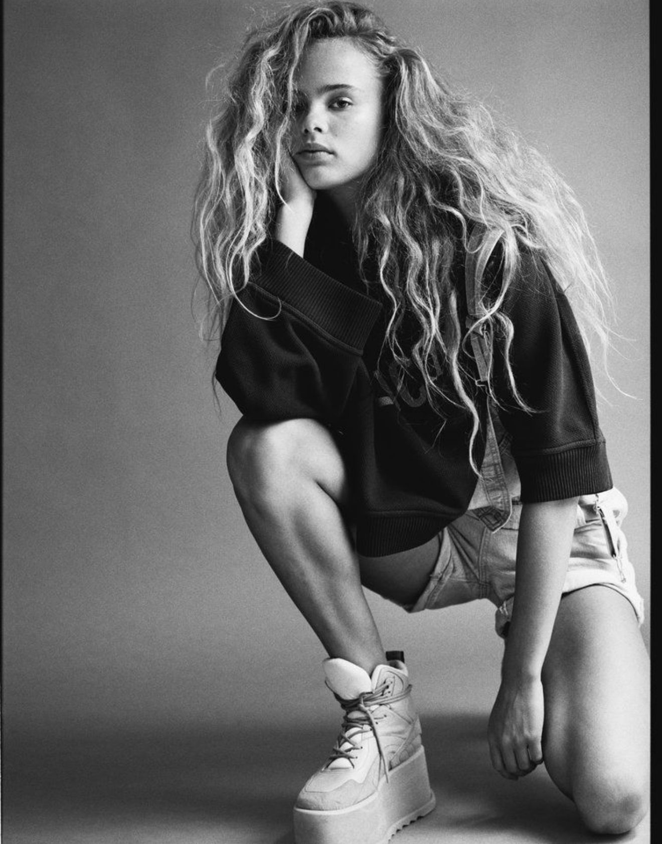 Young woman with long curly hair, squatting, wearing platform sneakers and a dark sweatshirt.