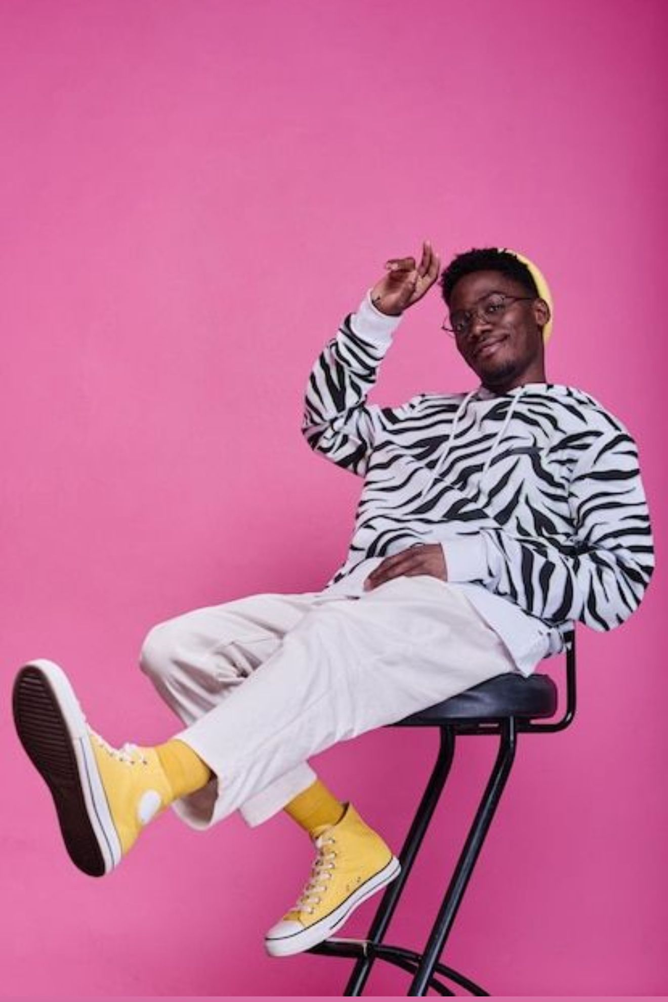 Man in zebra-striped sweatshirt, yellow shoes, and cap, sits on a stool, smiles at camera, pink background.