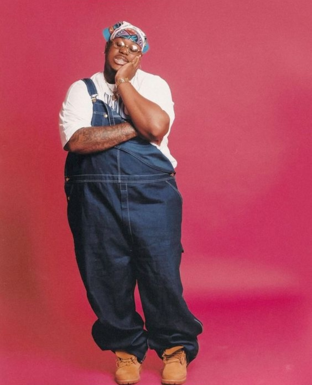 A person wearing denim overalls, white t-shirt, and a bandana leans against a pink backdrop with a relaxed expression.