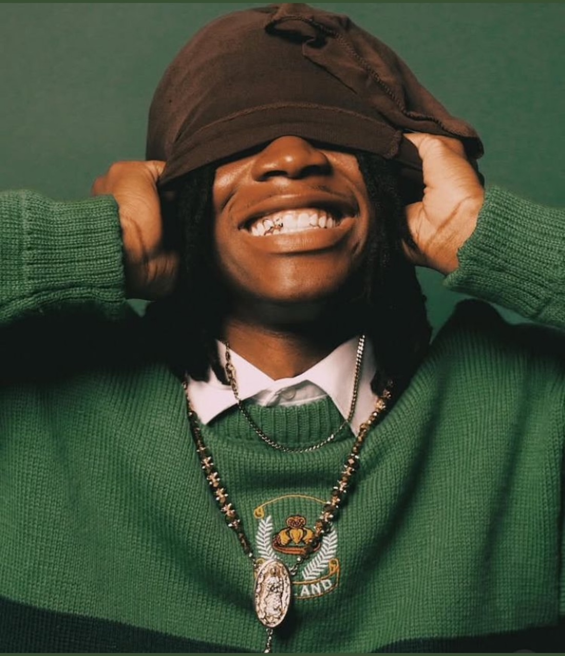 Person with long dark hair and wide smile pulling a brown hat over their eyes; wearing a green sweater and a beaded necklace.