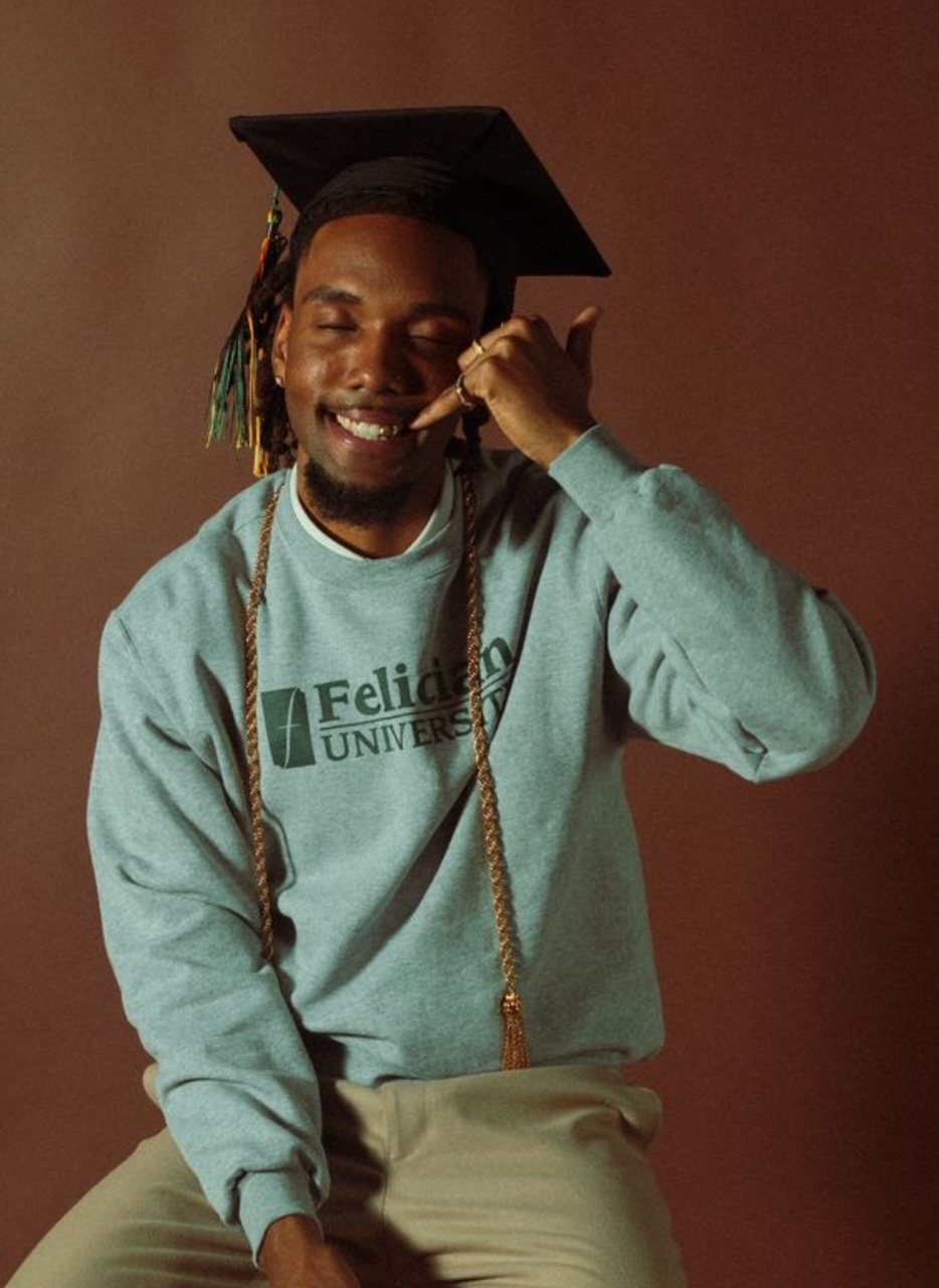 Man in graduation cap and Felician University sweatshirt makes phone gesture, smiling.