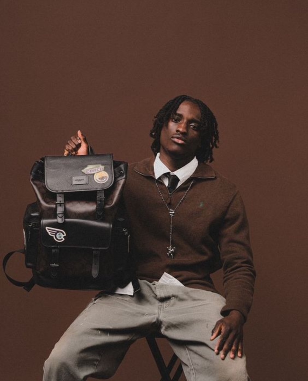 Man in brown outfit holding a brown backpack, sitting against a brown backdrop.