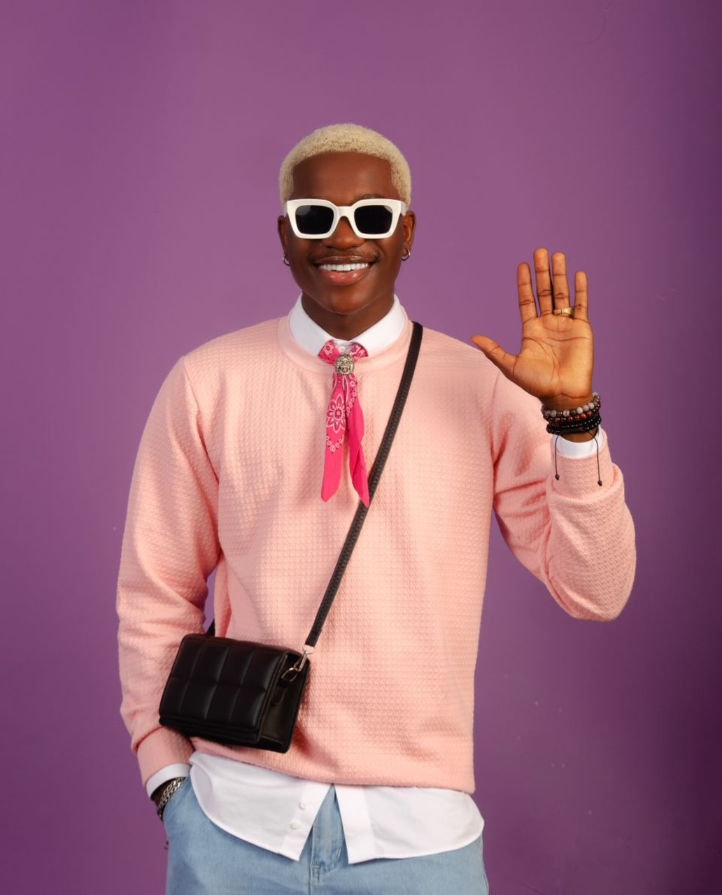 Smiling person with blonde hair, white sunglasses, and pink sweater waves, standing against a purple background.