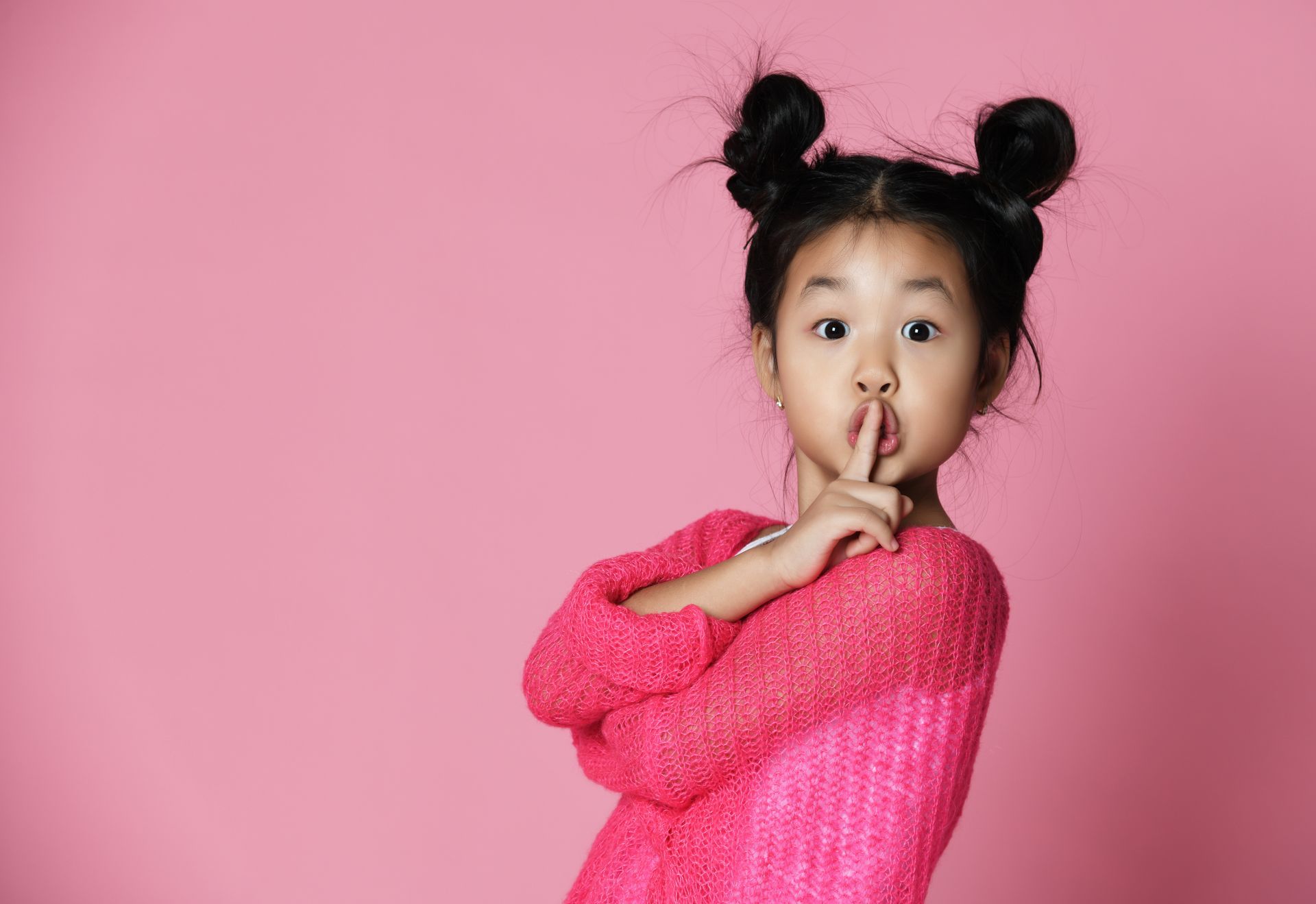 Girl with space buns, pink sweater, finger to lips, making a 