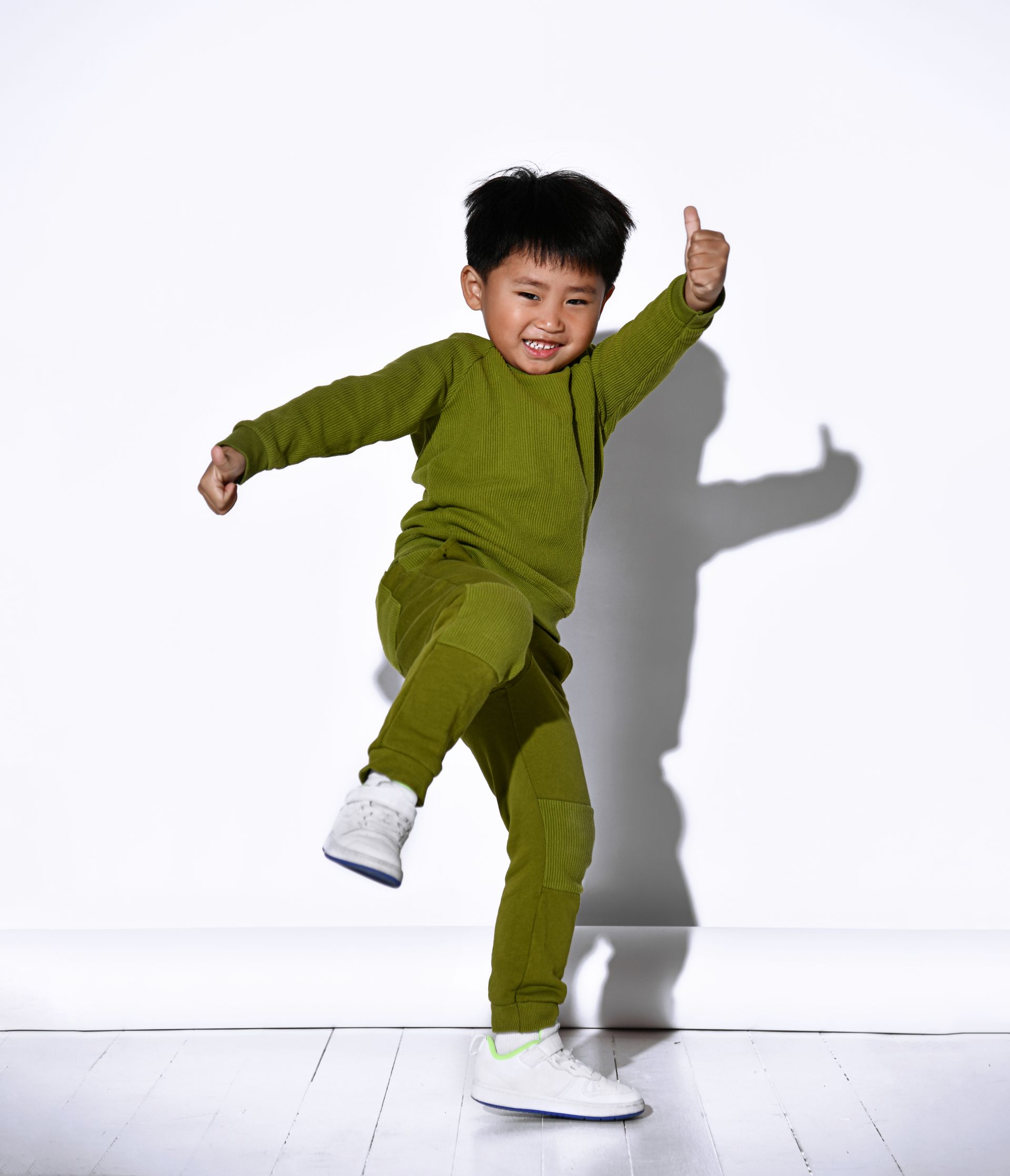 Young child in green outfit giving a thumbs-up and dancing. White background.