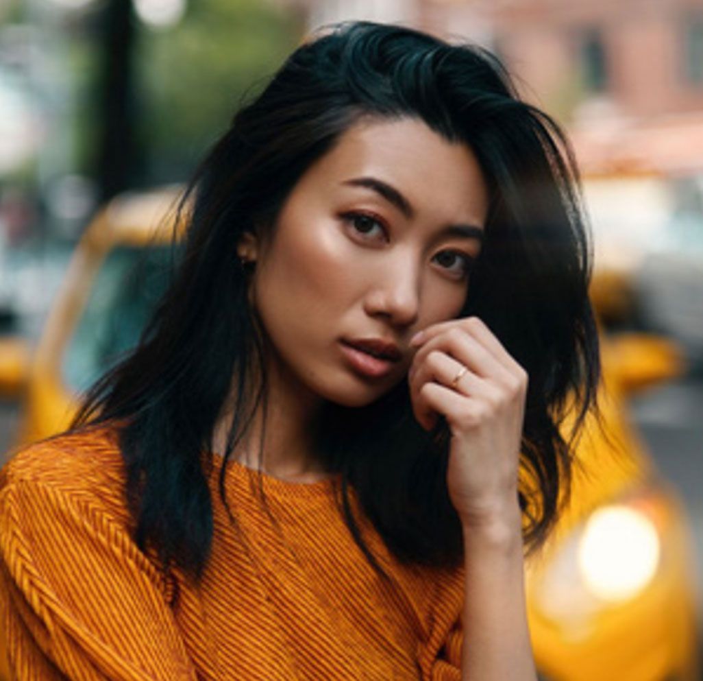 A woman in an orange sweater is standing in front of a yellow taxi