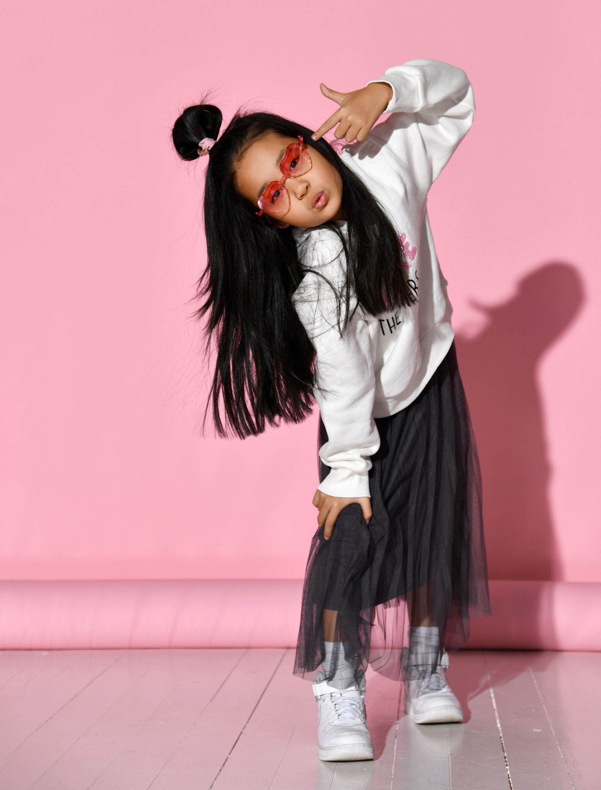 Young girl in sunglasses and white sweatshirt, making a gun hand gesture, leaning to the side. Pink background.