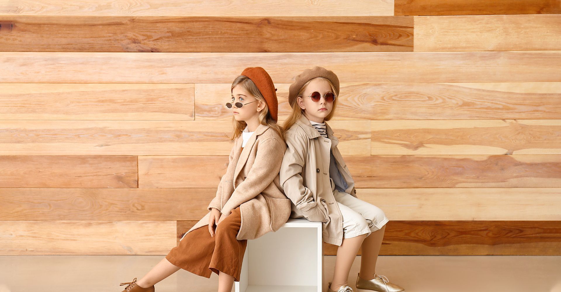 Two young girls in stylish outfits sit back-to-back on a white cube, against a wood-paneled wall.