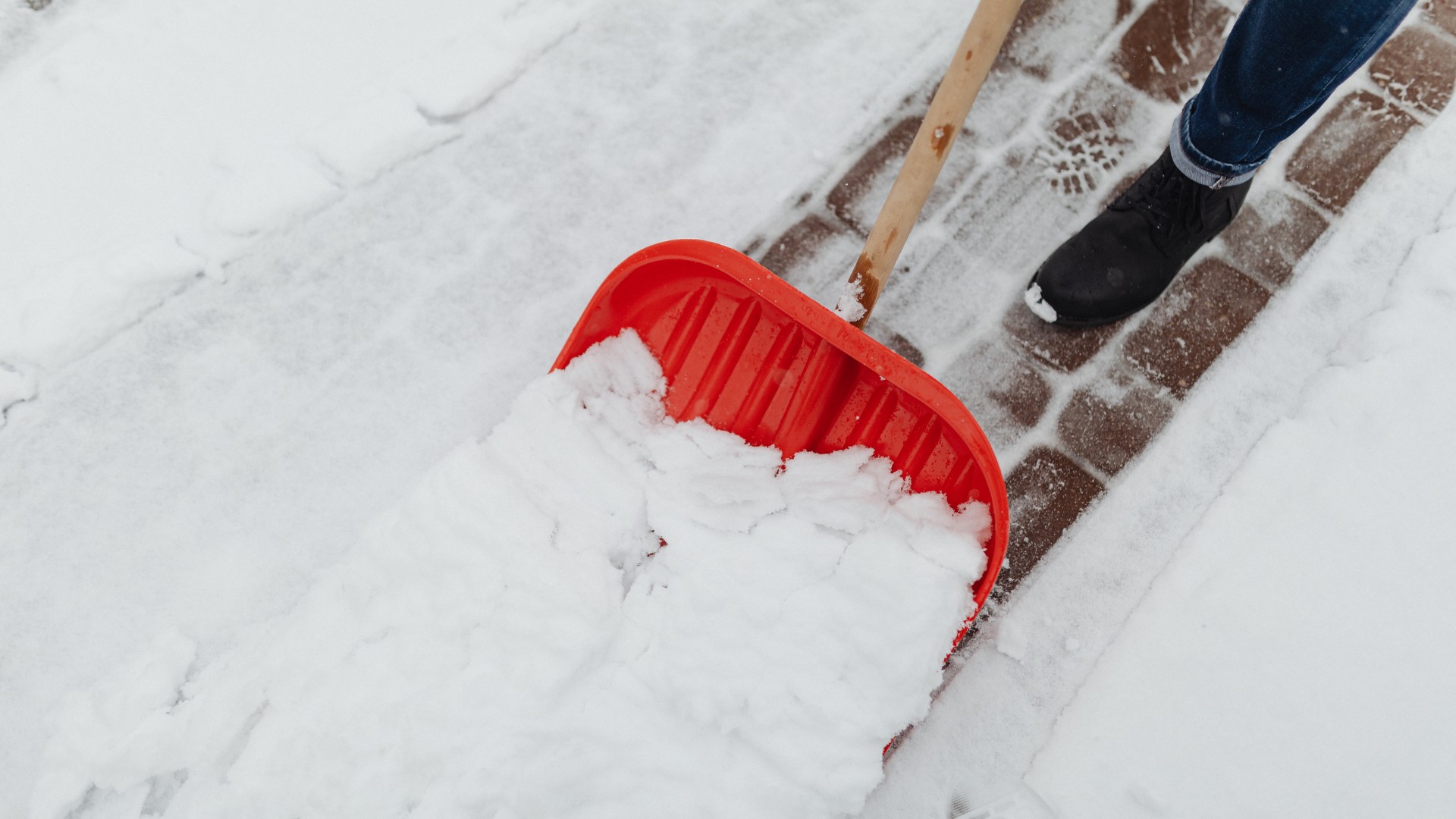 A person is shoveling snow from a sidewalk with a red shovel.