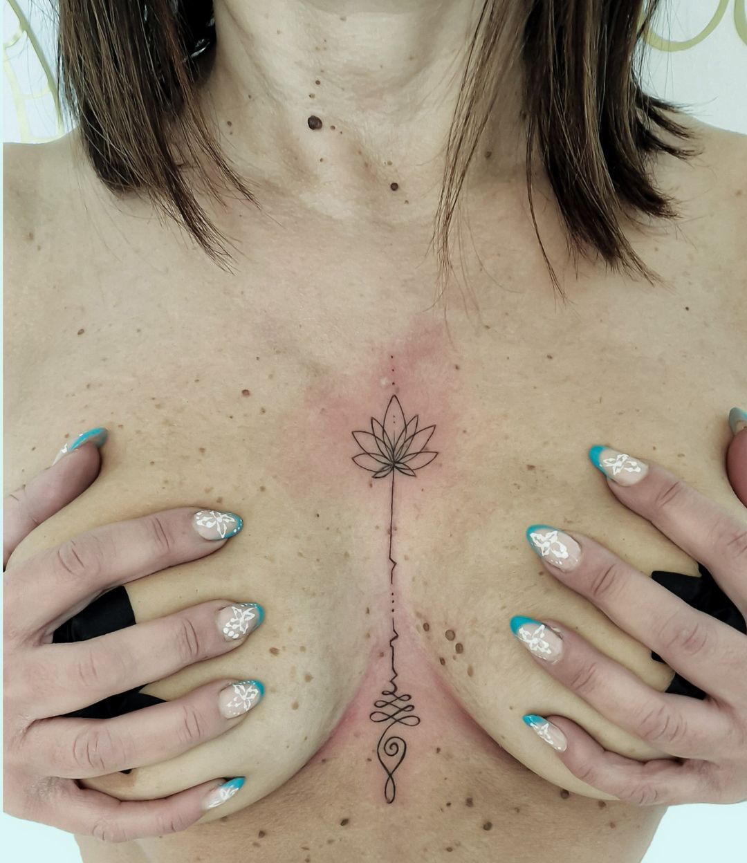 Petto di donna con un tatuaggio floreale verticale tra i seni, mani che incorniciano il disegno.
