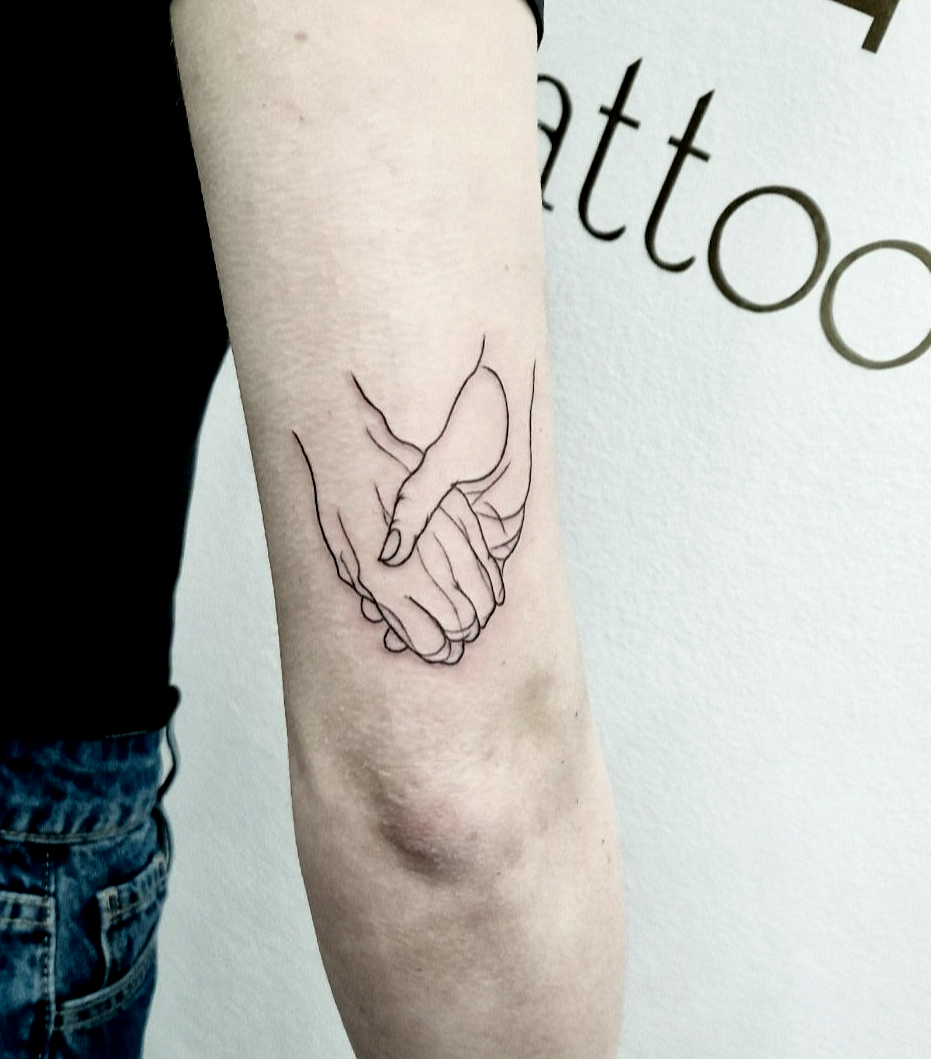 Primo piano di un avambraccio con un piccolo tatuaggio stilizzato nero raffigurante due mani che si stringono vicino al gomito.