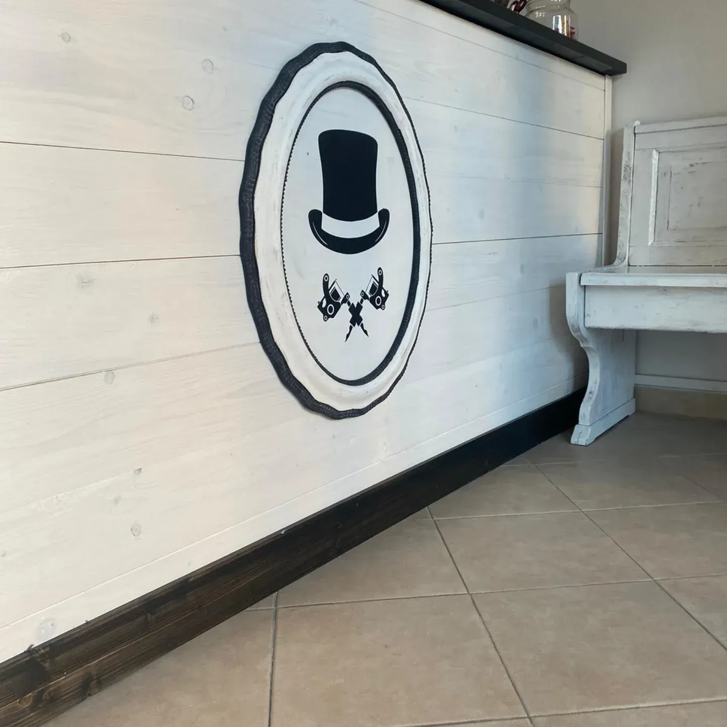 Parete bianca con logo nero raffigurante un cappello a cilindro e utensili incrociati accanto a un pianoforte bianco in una stanza piastrellata.
