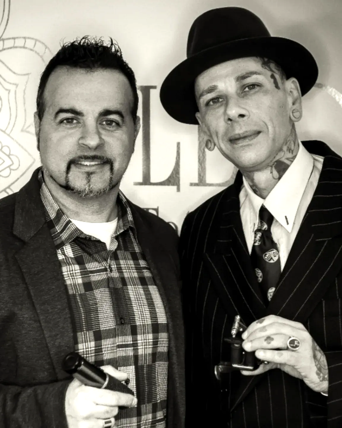Due uomini in abito e cappello posano insieme a un evento, con in mano dei sigari e un sorriso stampato in faccia.