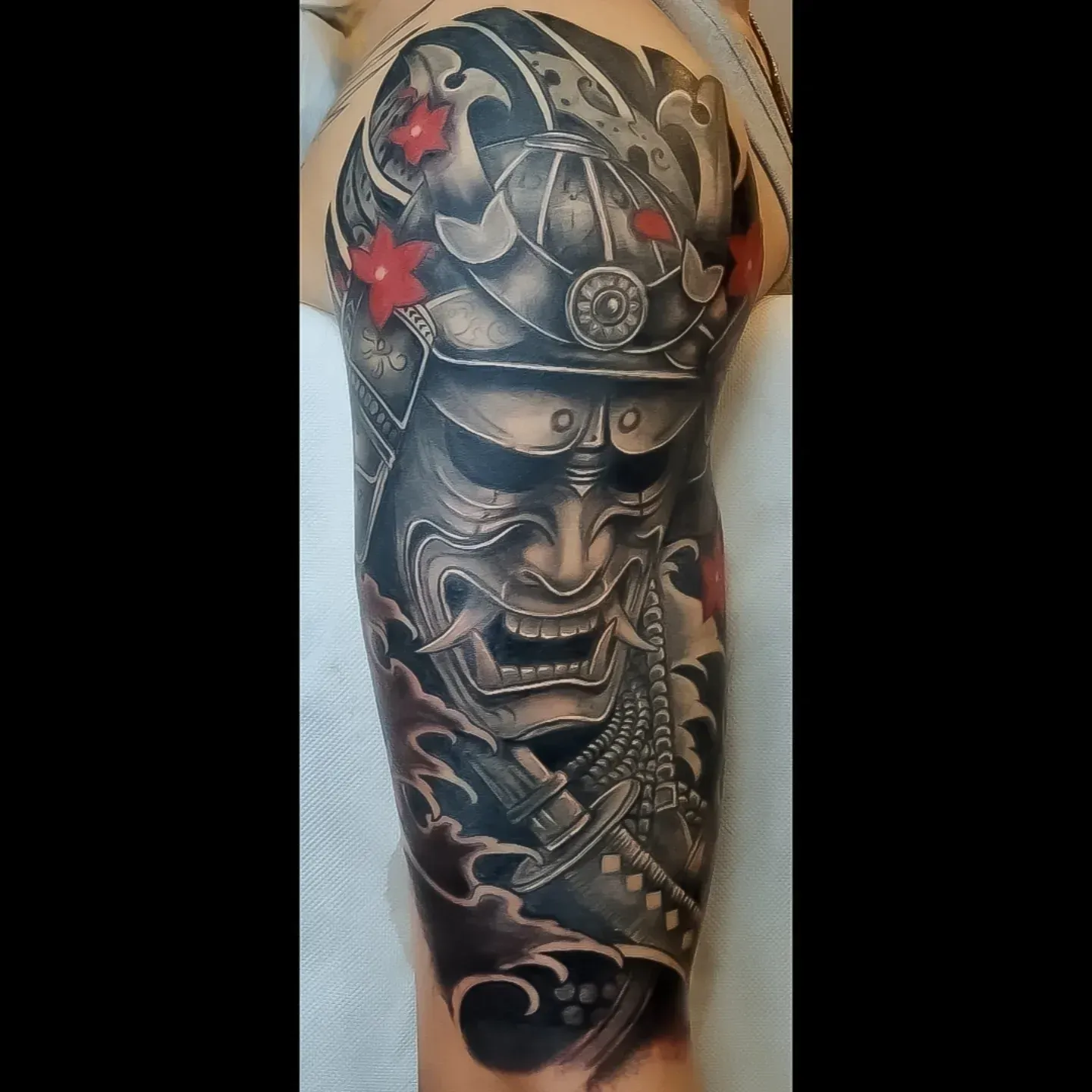 Tatuaggio in bianco e nero raffigurante un elmo da samurai e un volto fiero su un braccio, con dettagli rossi.