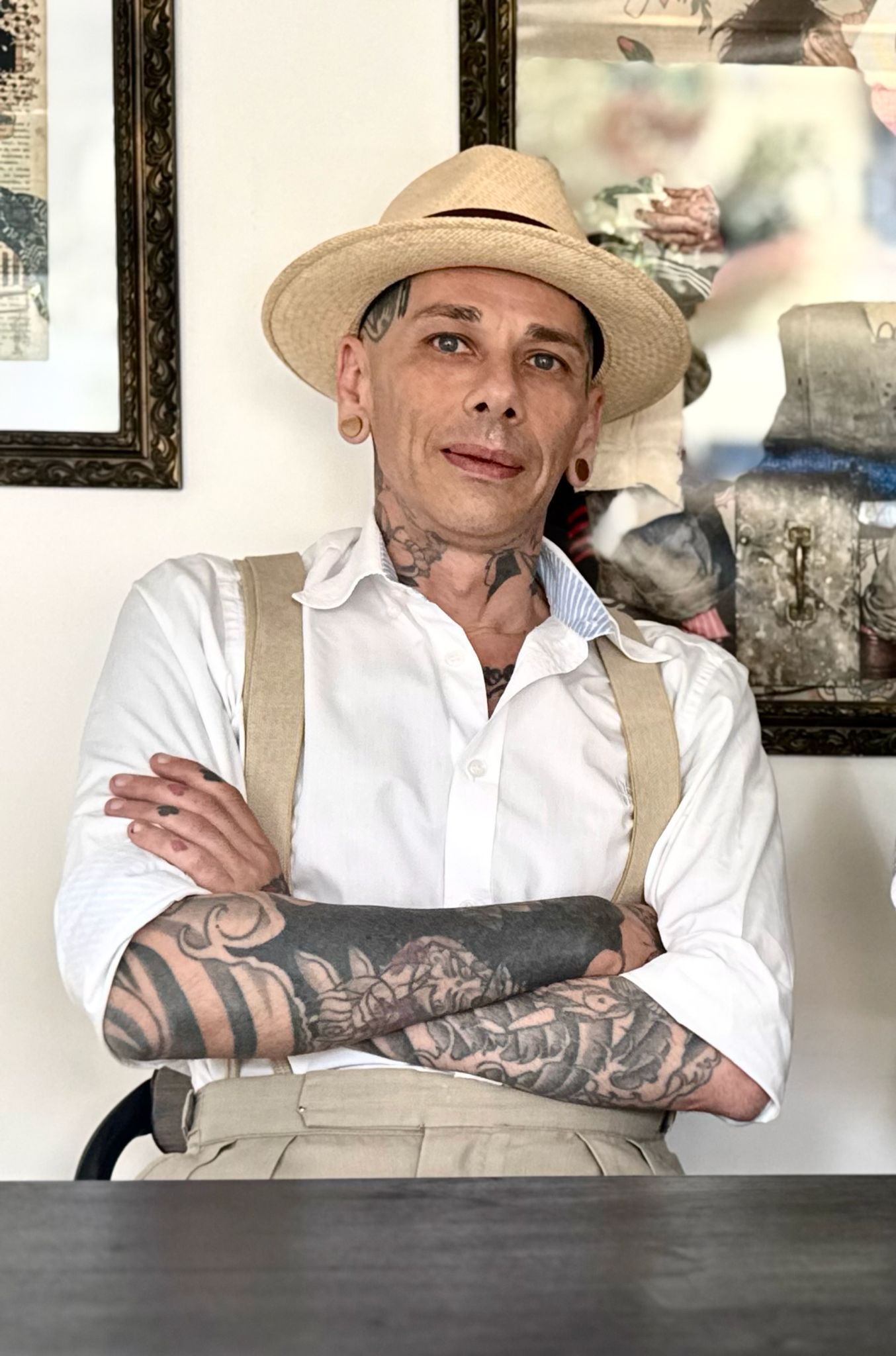 Persona con cappello di paglia e camicia bianca, con braccia tatuate, in piedi in un ambiente chiuso con le braccia incrociate.