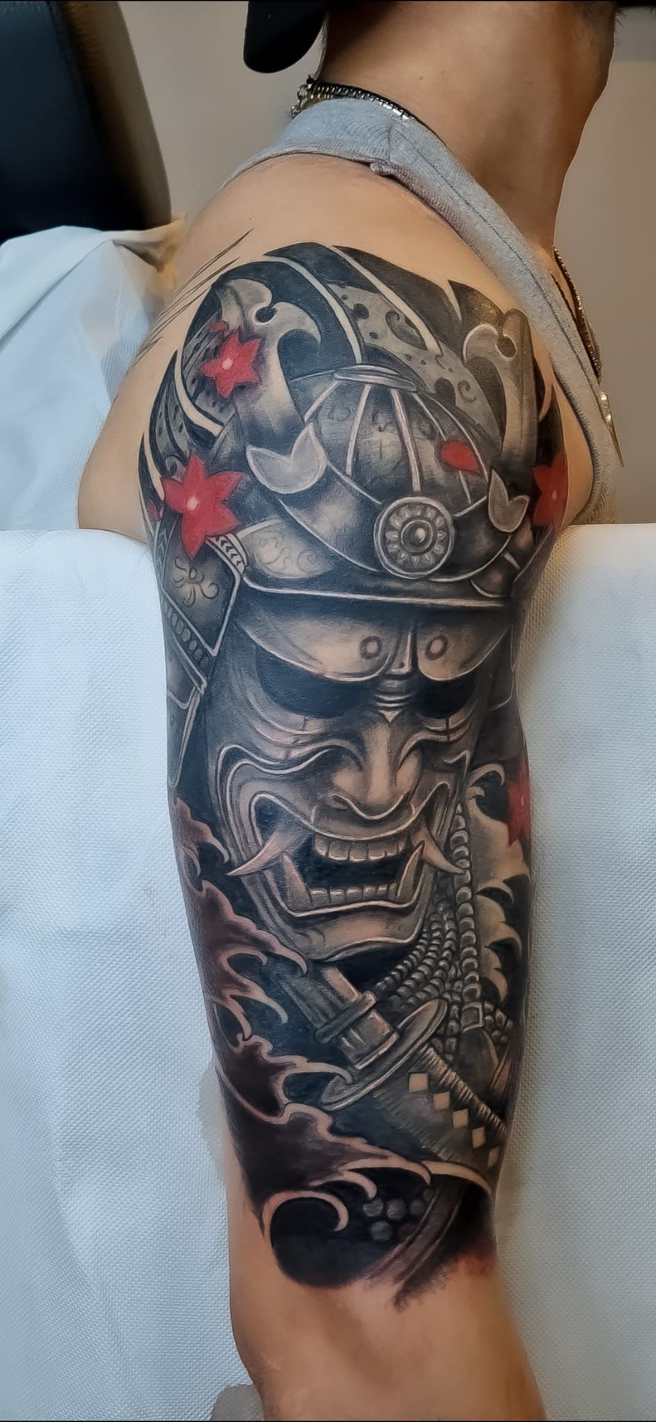 Primo piano di un braccio con un tatuaggio dettagliato raffigurante il volto di un samurai, realizzato in nero, grigio e rosso.