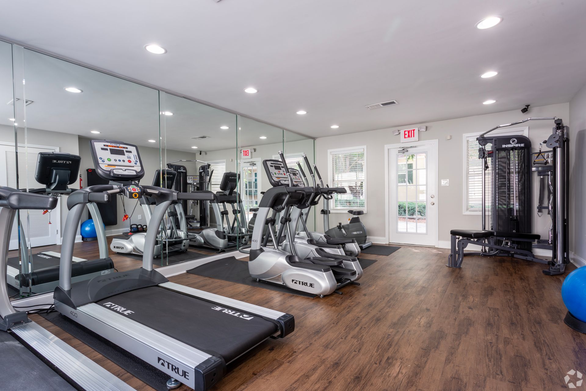 A gym room with wood floors, featuring three treadmills in front of a mirrored wall and a multi-station weight machine.