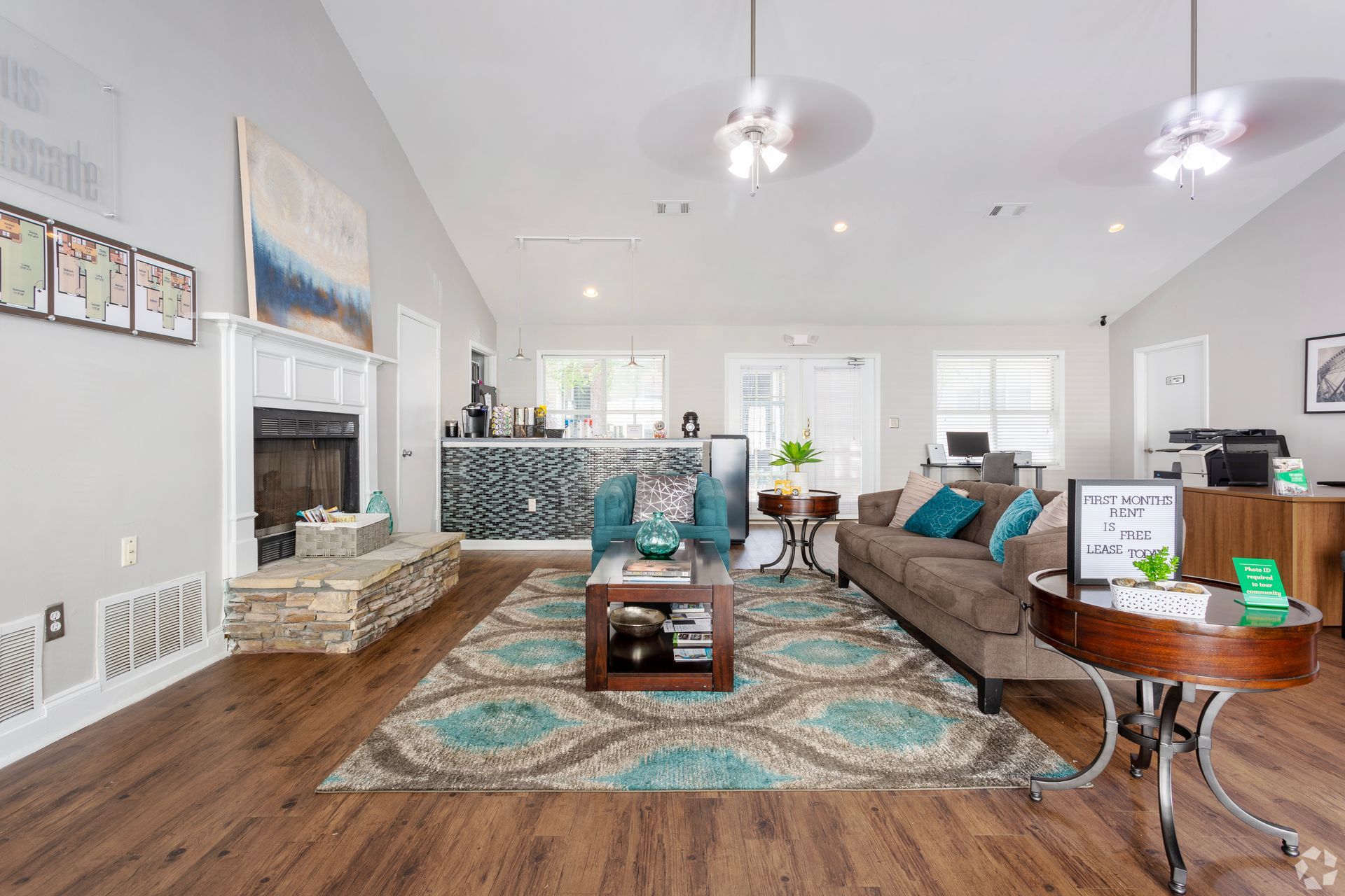 A spacious, brightly lit community lounge with wood floors, a fireplace, teal accents, and a brown sofa on an area rug.