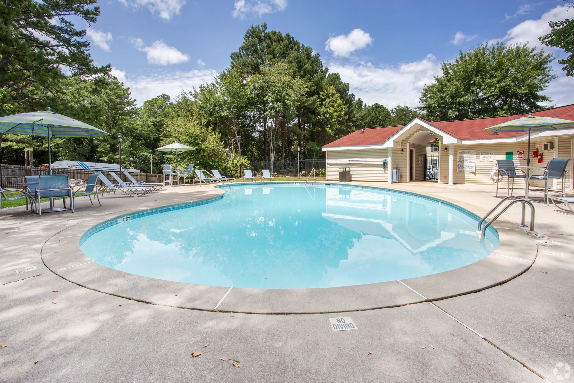 A turquoise swimming pool surrounded by a concrete deck, lounge chairs, umbrellas, and a small building under a blue sky.