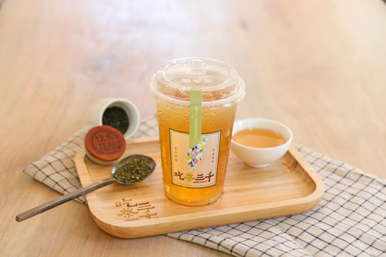 Iced fruit tea in a clear cup with a green straw on a wooden tray, with tea scoops and a small sauce cup.