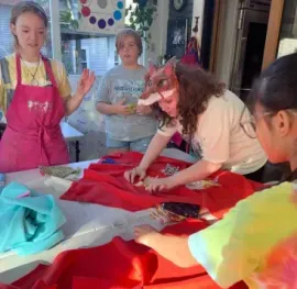 Four people work together around a table covered in red fabric and sewing supplies in a bright, indoor classroom.