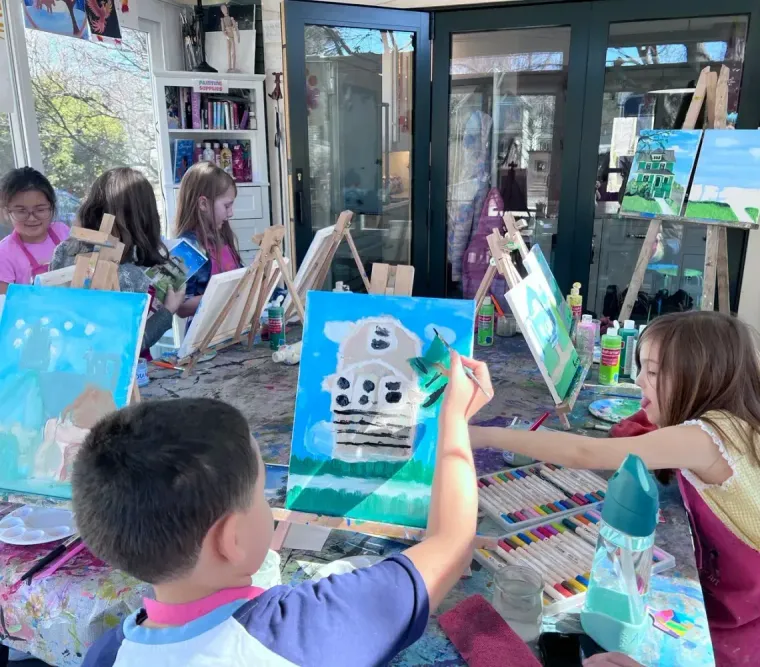 Children sit at tables in a bright, sunlit room, painting landscapes on canvases set on wooden easels.