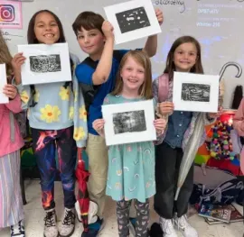 Six children stand in a classroom, smiling and holding up their black and white art prints for the camera.