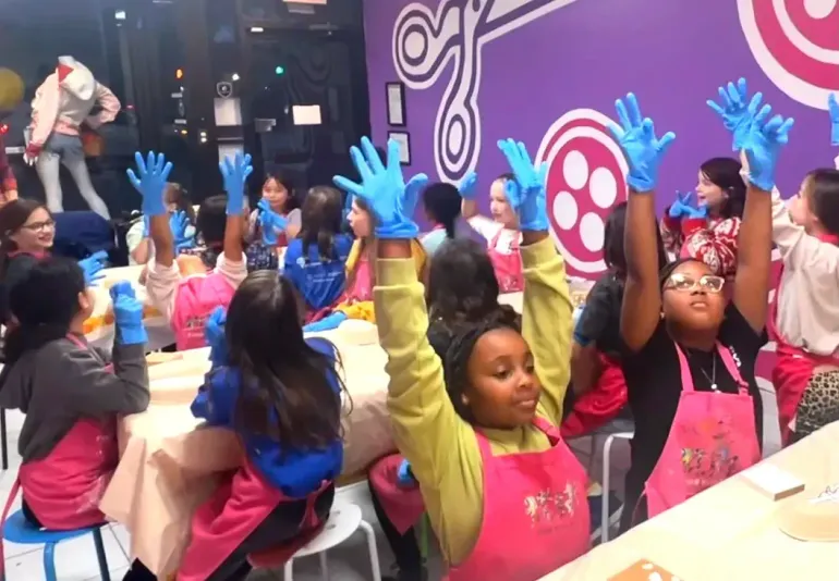A group of children wearing pink aprons and blue plastic gloves sit at tables in a purple room, hands raised in the air.