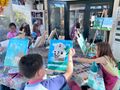 Three children with paint-covered hands pose with outstretched arms in a bright art studio filled with supplies.