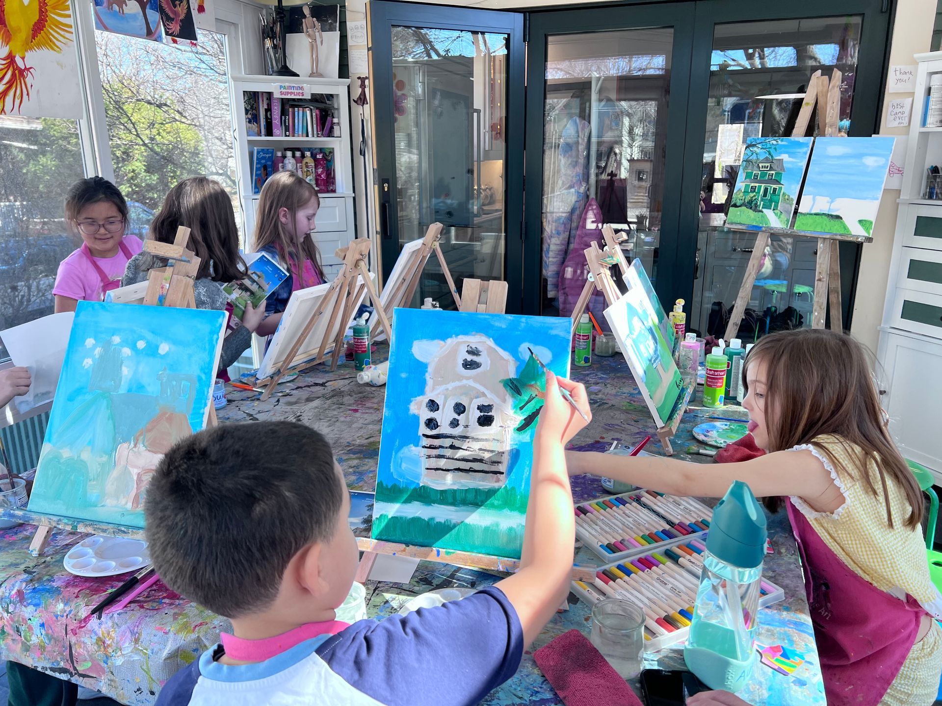 Three children with paint-covered hands pose with outstretched arms in a bright art studio filled with supplies.
