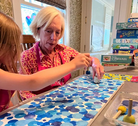 A person and a child work together to paint a long board with blue circles using sponge dabbers at a table.