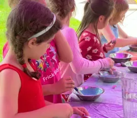 Four children participate in an outdoor art activity, painting with small bowls of blue paint at a purple-covered table.