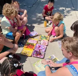 A group of children and an adult sit on a sidewalk, sorting colorful craft supplies into small plastic trays.