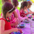 Four children in red and pink shirts paint at a purple table outdoors, each working with a small metal bowl of paint.