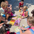 A group of children and an adult sit on a sidewalk, sorting colorful craft beads into small trays.