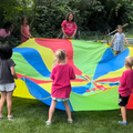 A group of children plays with a colorful, multi-paneled circular parachute in a grassy park setting.