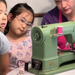 An adult in a purple shirt and pink apron demonstrates how to use a vintage green sewing machine to two attentive children.