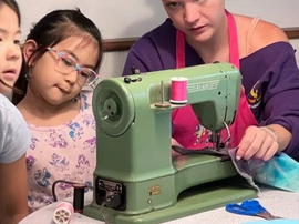 A person uses a vintage green sewing machine to stitch fabric while two children watch with focused interest.