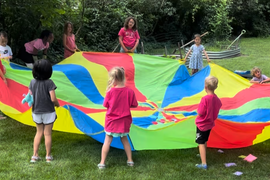 Children standing in a circle outdoors, holding the edges of a colorful parachute as they play a game on the grass.