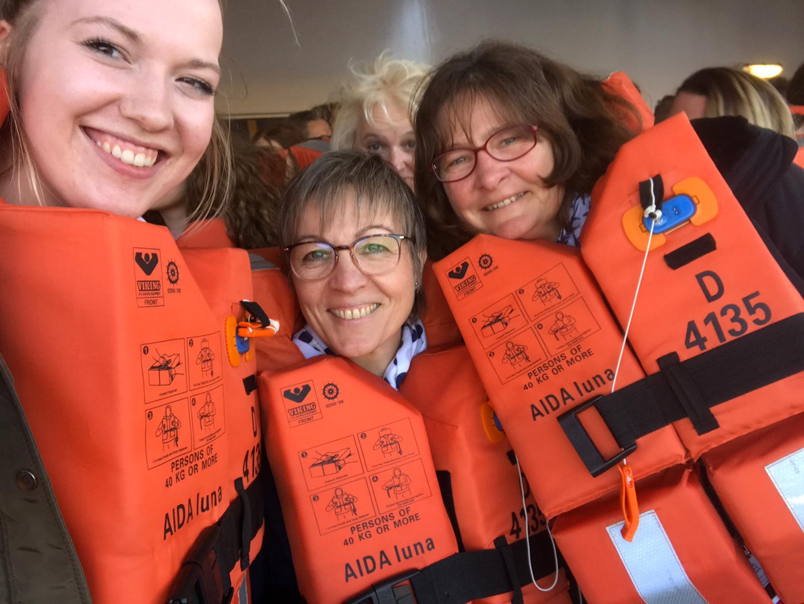 Drei lächelnde Personen in leuchtend orangefarbenen Schwimmwesten posieren eng beieinander in einem Innenraum.
