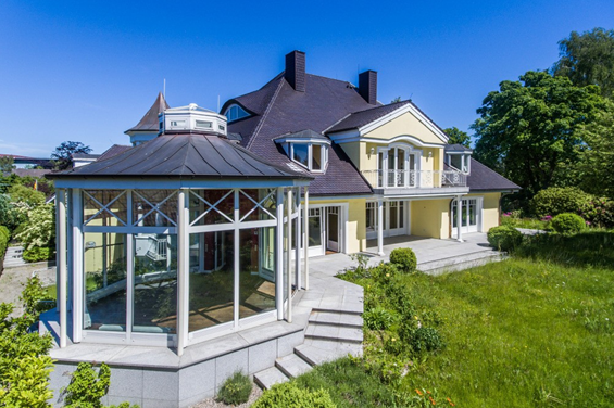 Elegantes cremefarbenes Haus mit dunklem Dach und gläsernem Pavillon mit Blick auf einen grünen Rasen unter blauem Himmel