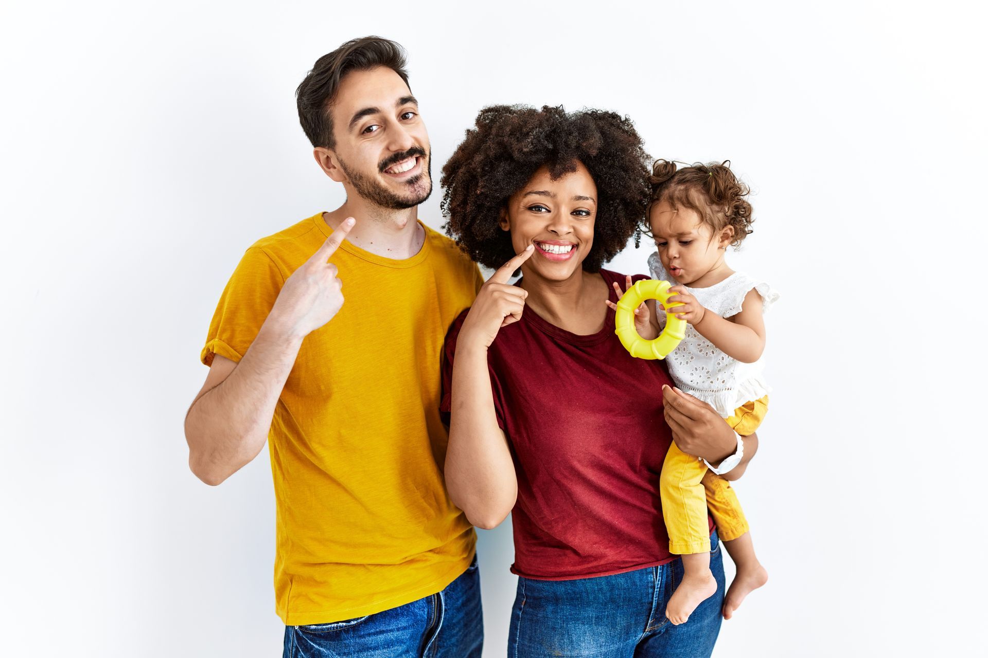 Happy parents and toddler pointing to their healthy smiles at a family dentist appointment.