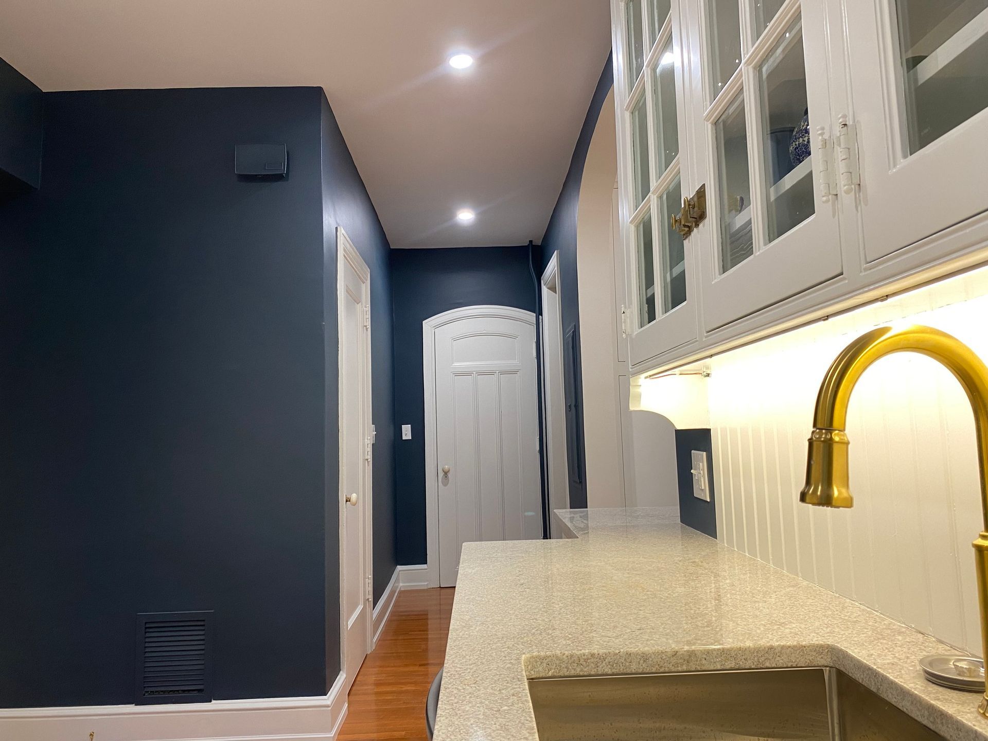 Narrow hallway with dark blue walls, white door, wood floor, and a bright kitchen counter with brass faucet.