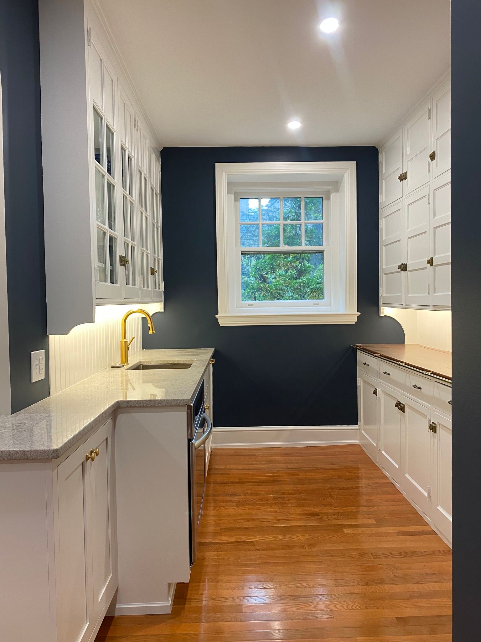 Narrow galley kitchen with white cabinets, gold faucet, dark blue walls, and a window at the far end.