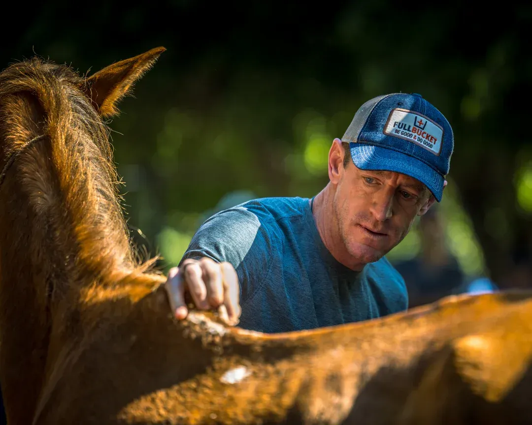 Man petting a brown horse outdoors; man wears a blue cap, and the horse is the focus.